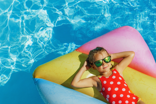 Kind mit Sonnenbrille liegt auf Luftmatratze im Pool und verschr&auml;nkt die Arme hinter dem Kopf